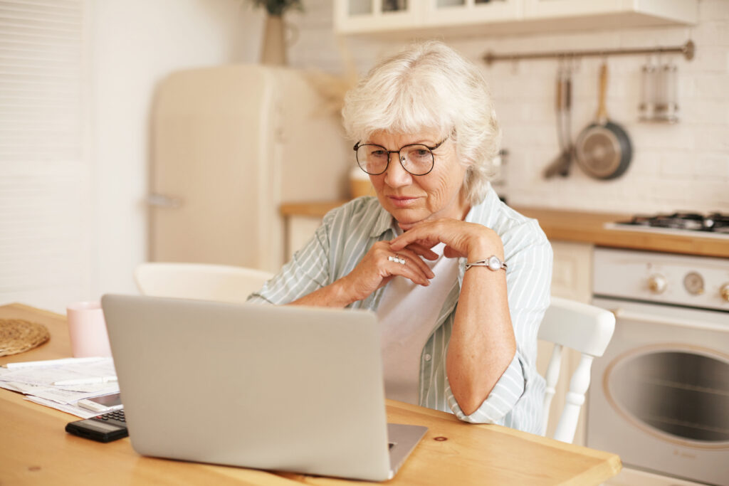 nowoczesna technologia ludzie starsi i emerytura siwa emerytka w okularach wypelniajaca formularz wniosku o pozyczke online siedzaca przed laptopem czytajaca informacje z powaznym skupionym spojrzeniem - Uniwersytet Trzeciego Wieku Online