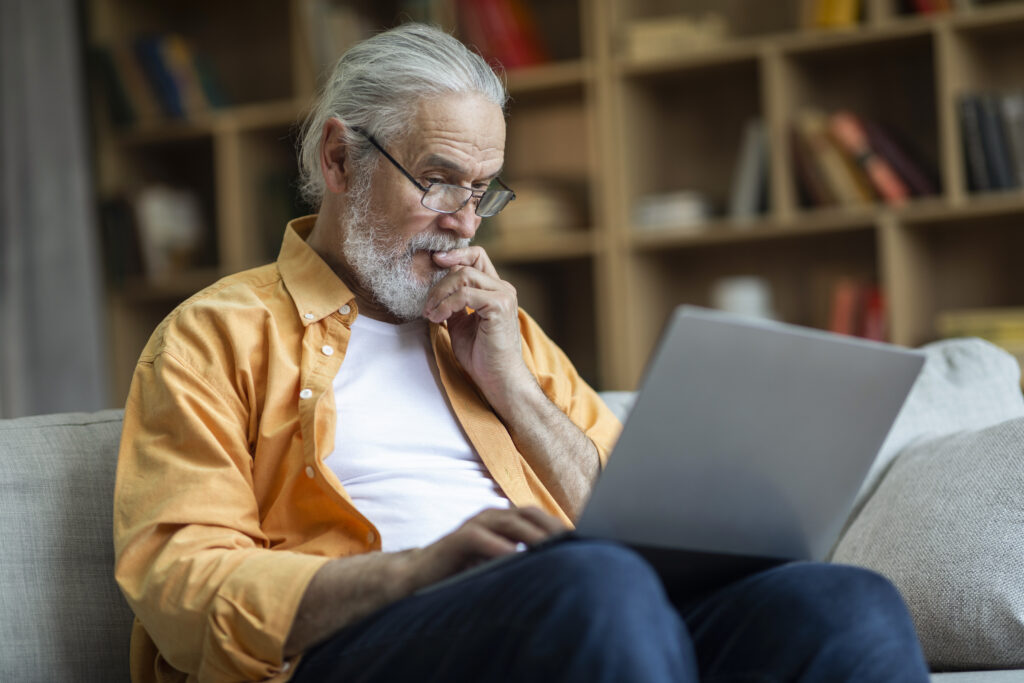 zamyslony dziadek surfuje po sieci podczas odpoczynku w domu przy laptopie - Uniwersytet Trzeciego Wieku Online